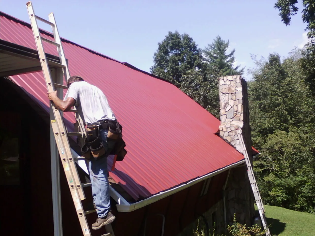 Roofer on a ladder beside a red metal roof and stone chimney — Soffit & Fascia Repair by Roofing Brothers in Coeymans Hollow