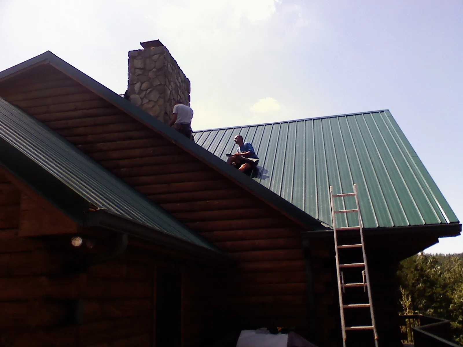 Roofers installing green metal panels beside a stone chimney on a log home in Coeymans Hollow