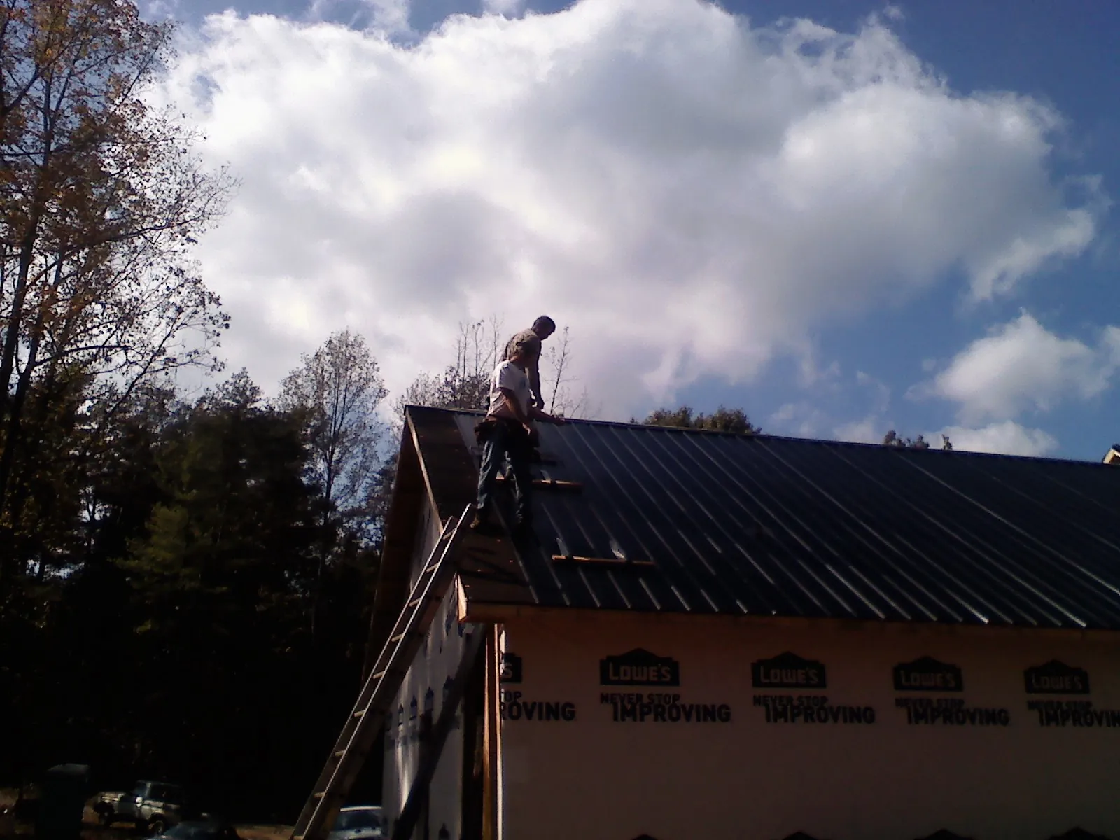 Roofer fastening dark metal panels high against a cloudy sky, from a Roof Inspection job in Coeymans Hollow